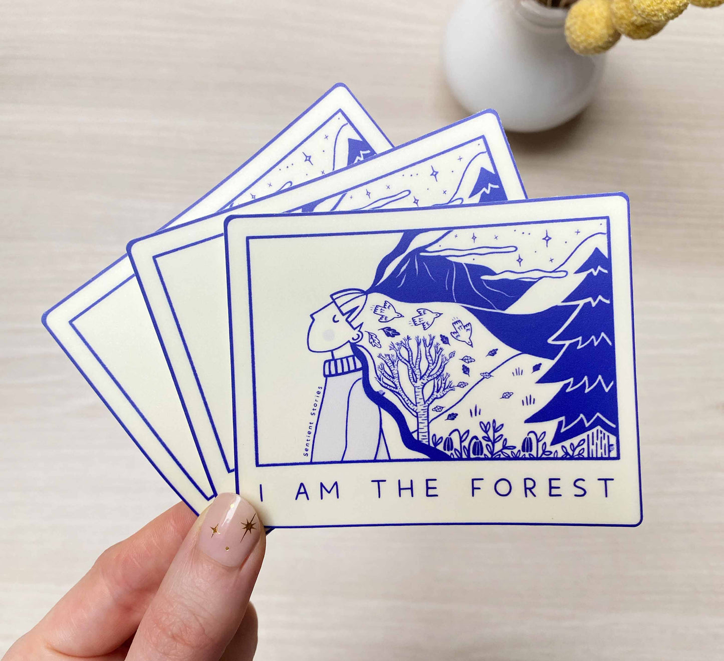 A hand holding several blue and white vinyl stickers with a nature-themed illustration, including trees and mountains, with the text 'I AM THE FOREST' on one of them.