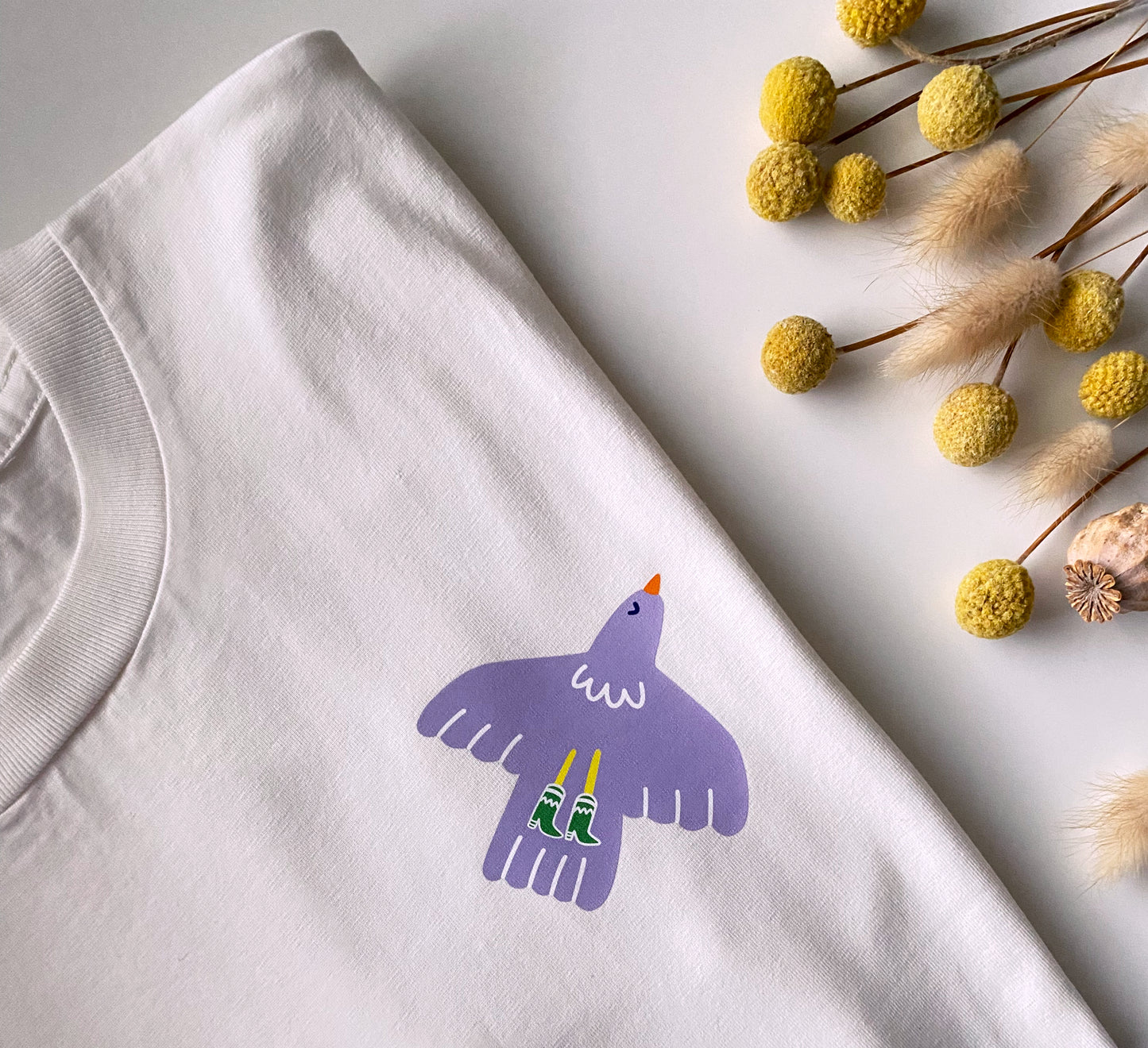 White t-shirt with a purple bird graphic on a light gray surface with dried flowers.