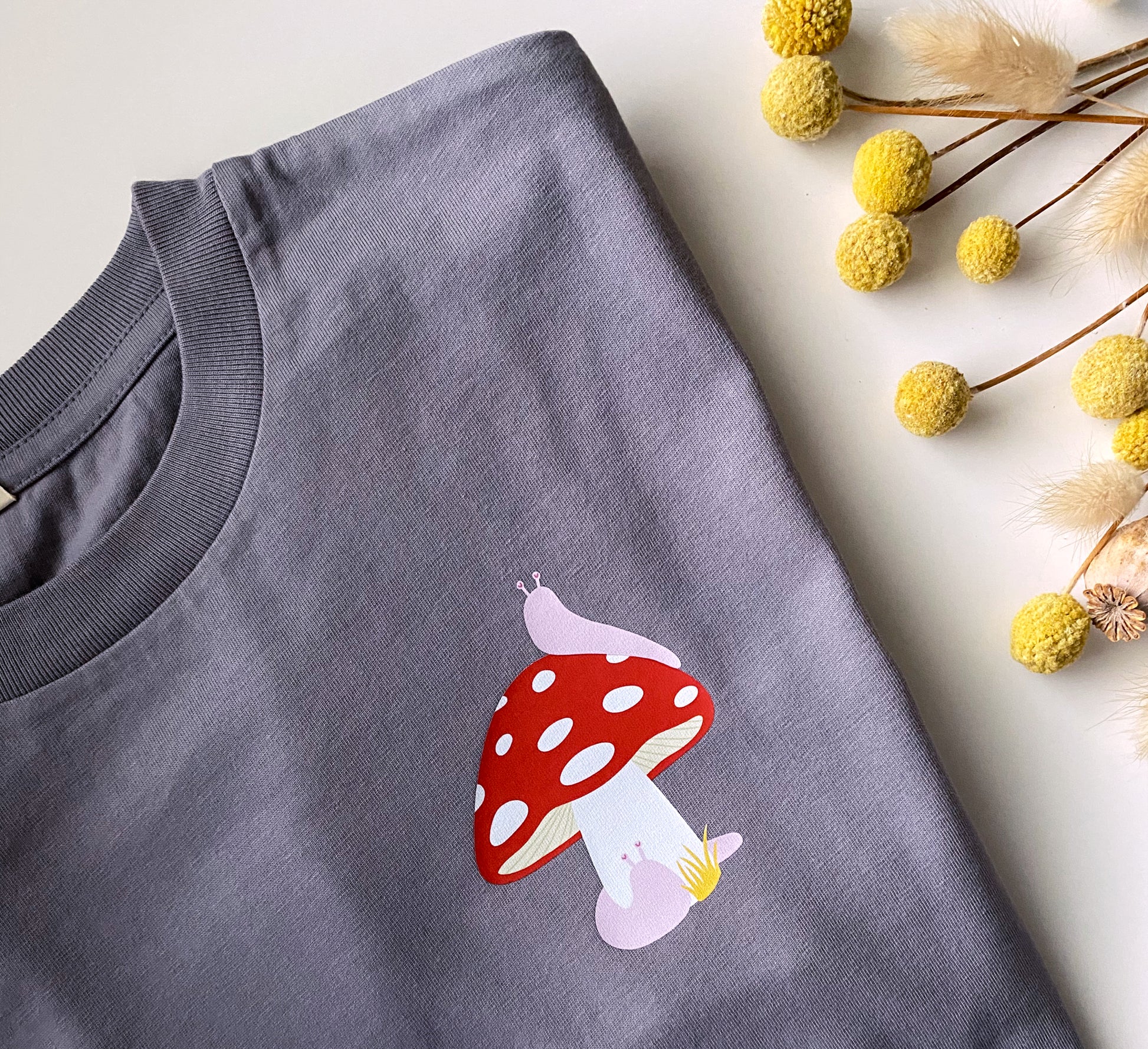 Gray t-shirt with a red and white mushroom design on a light surface with yellow flowers.