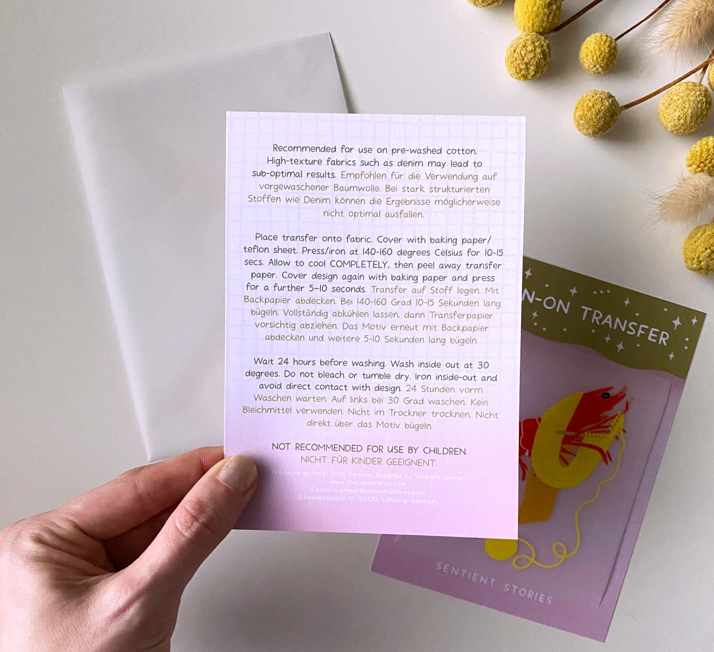 Hand holding a printed sheet with text, surrounded by fabric transfers and flowers on a light surface.