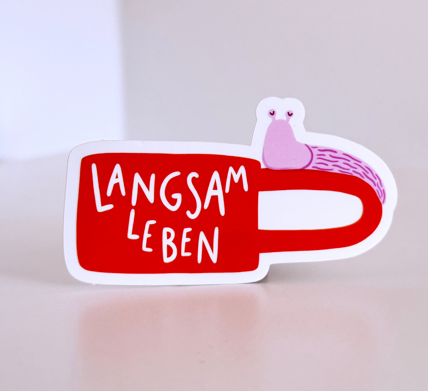 A die-cut sticker shaped like an orange coffee mug with the German phrase 'LANGSAM LEBEN' in white lettering and a small pink snail illustration on the handle, shown on a light beige surface against a neutral background.